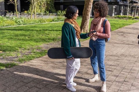 Asian and african american college friends talking outdoors with skateboard and smartphone