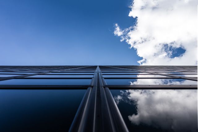 Upward view of a modern glass skyscraper reflecting blue sky and clouds. Ideal for business, corporate, and architectural themes, representing ambition, growth, and urbanism in marketing and promotional materials.