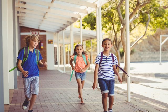 Three cheerful school children running in a school corridor with backpacks on a sunny day. Ideal for educational materials, back-to-school promotions, and advertisements focusing on childhood joy and learning.
