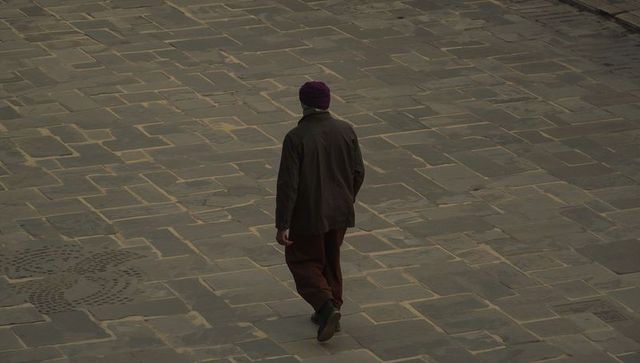 Solitary man walking across empty stone plaza wearing purple beanie under overcast sky, moody quiet