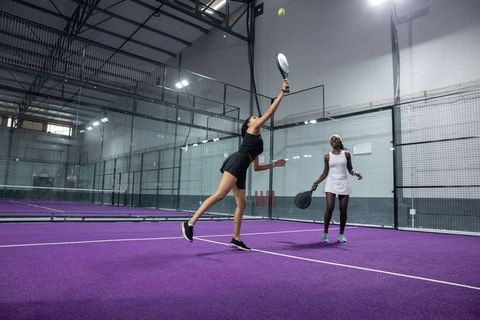 Women playing padel on vibrant purple indoor court, reaching for overhead shot