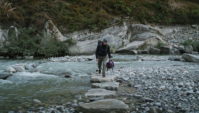 Solo hiker crossing river on stepping stones carrying backpack and purple duffel
