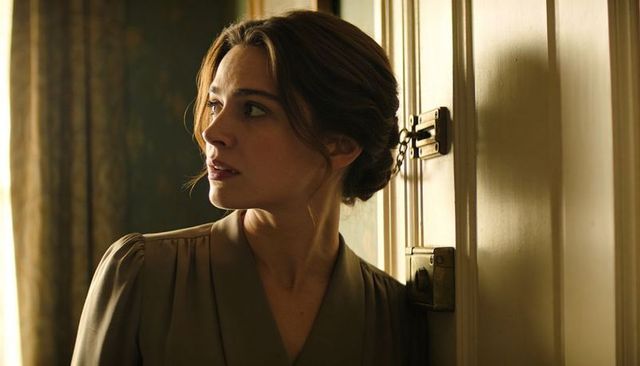 Tense woman listening beside locked door, vintage blouse and low bun, cinematic sidelight