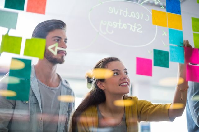 Smiling executives writing on sticky notes on glass wall in office