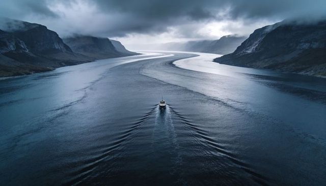 Boat gliding through s-shaped fjord cutting v-wake between rocky cliffs under moody clouds