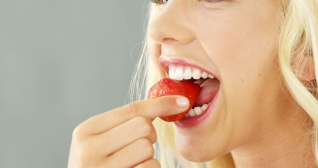 This close-up image of a blonde woman eating a fresh strawberry shows her smiling and enjoying a healthy snack. Ideal for use in articles, advertisements, and blogs promoting healthy eating, nutrition, wellness, fresh produce, dieting tips, dental health, and overall well-being.