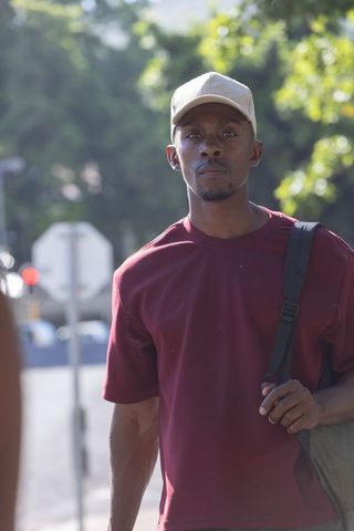 African american man walking on sunlit street wearing cap and carrying shoulder bag