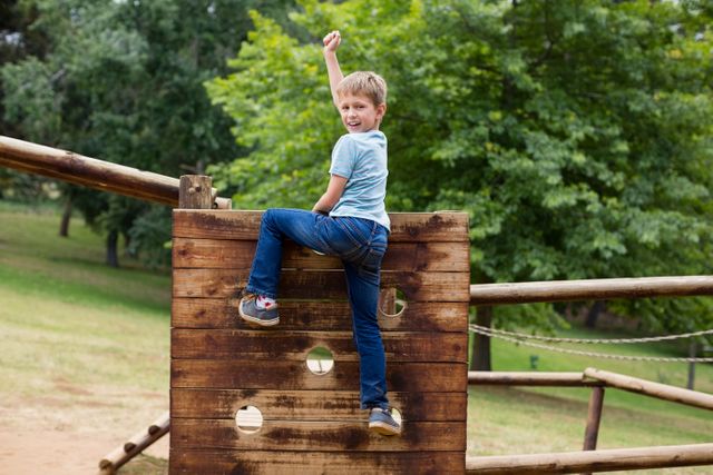 Boy climbing a wooden playground structure in a park on a sunny day. Ideal for use in articles or advertisements related to outdoor activities, children's play, healthy lifestyles, and family outings. Perfect for promoting parks, playgrounds, and recreational areas.