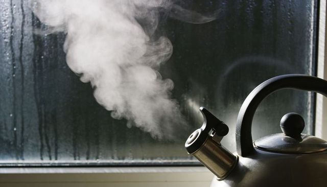 Stainless steel kettle releasing steam by foggy window, whistle spout and dark handle