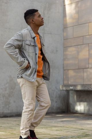 African american man standing by urban tiled wall wearing denim jacket and orange sweater