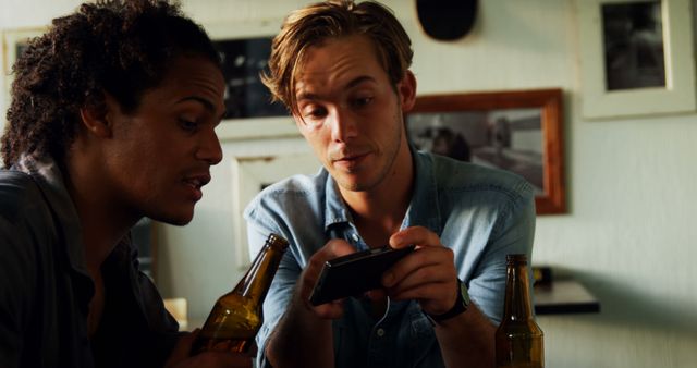 Two friends sitting together at home, engaging with a smartphone while enjoying bottles of beer. One person is focused on the phone, showing something of interest to the other. Casual atmosphere, perfect for themes of friendship, leisure, technology, and modern lifestyle. Useful for advertising social apps, home decor, or friendly gatherings.