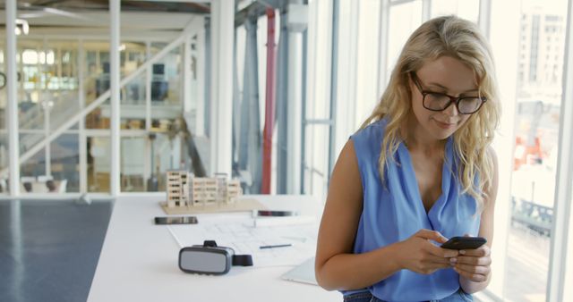 This image features a young professional woman using a smartphone in a modern office space. She is wearing glasses and a blue blouse, suggesting a casual yet professional work environment. This image is ideal for use in business and technology related content, articles about modern offices, communication technology, or women in business.