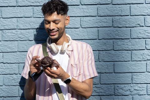 Young indian man smiling while enjoying chocolate muffin against blue brick wall