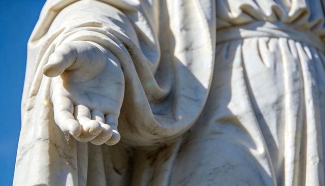 Sunlit marble statue holding outstretched hand with draped robes, close-up detail