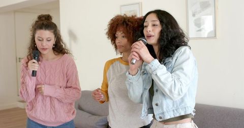 Diverse female friends singing karaoke at home holding microphones in cozy living room