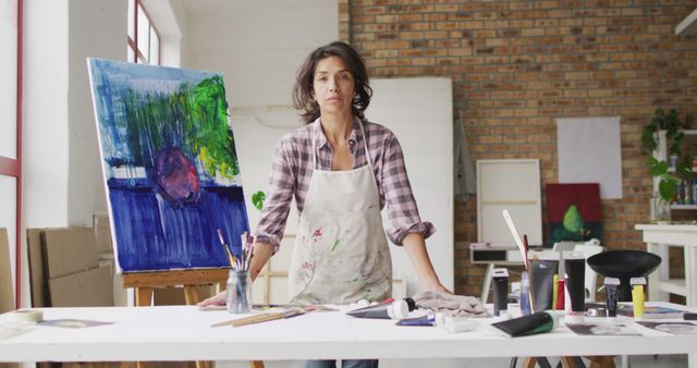 Female artist standing at work table with various art supplies in an art studio with brick walls, looking directly at the camera. Perfect for themes related to creativity, artistic process, women in art, and studio workspace.