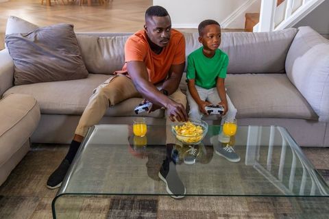 African american father and son playing video games on couch with snacks and juice