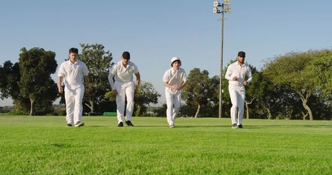Four cricketers walking across green field in white uniforms before match on sunny day