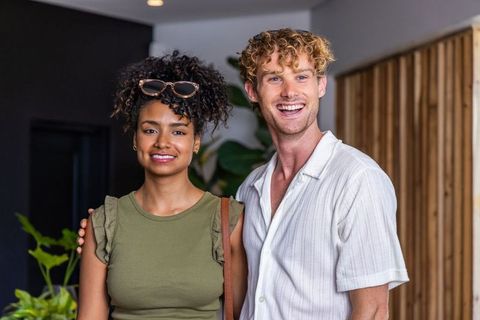Smiling diverse couple in modern lobby with arm around partner, casual urban portrait
