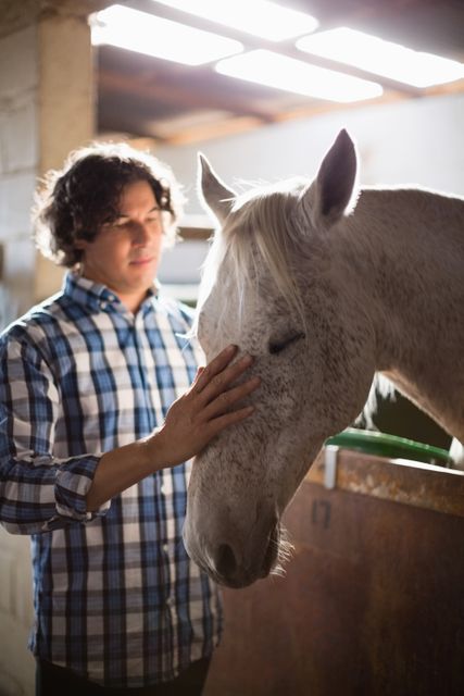 Man gently caressing horse in stable, showcasing bond between human and animal. Ideal for themes of animal care, equestrian activities, friendship, and gentle interaction. Suitable for use in articles, blogs, and advertisements related to horse care, animal therapy, and rural lifestyle.