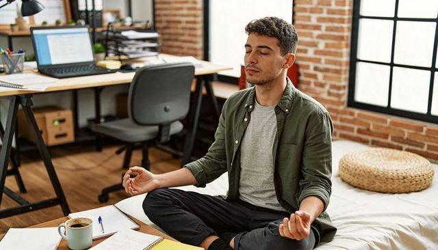 Meditating man finding focus on bed in loft workspace with laptop and notebooks