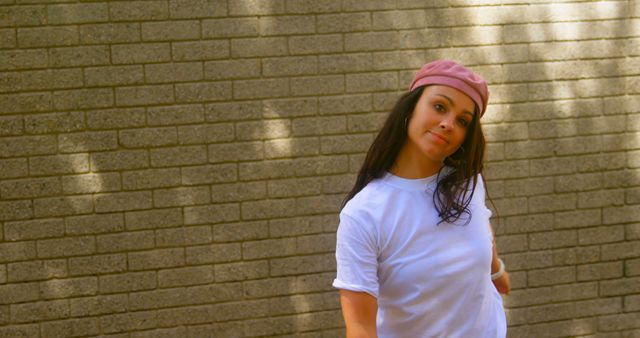 A young Caucasian woman stands confidently against a brick wall, wearing a pink headband and a casual white t-shirt, with copy space. Her relaxed posture and direct gaze convey a sense of ease and self-assurance.