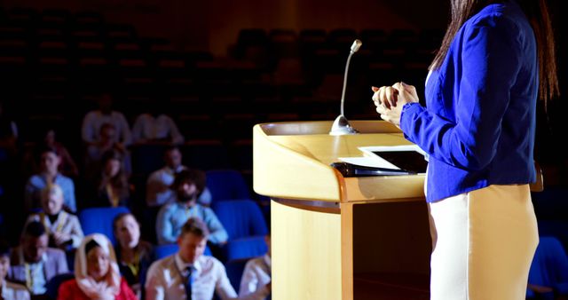 Businesswoman wearing blue jacket and white skirt giving presentation to audience in large auditorium. Image suitable for business conferences, seminars, social media posts on public speaking, professional interaction settings, meeting preparations, employee workshops, company events, or educational material.