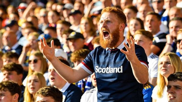 Red-haired bearded fan shouting and cheering passionately in scotland jersey at packed stadium