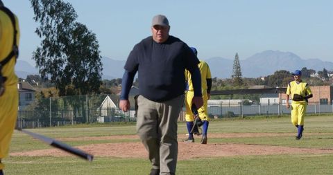 Coach walking off pitching mound with teammates in yellow uniforms on local baseball field