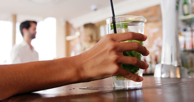 Person holding a glass of mojito with mint leaves at a bar counter. The background includes blurred individuals likely socializing. Useful for themes related to nightlife, social gatherings, alcoholic beverages, and leisure activities. Ideal for articles, advertisements, or blog posts about socializing, cocktail recipes, or bar environments.