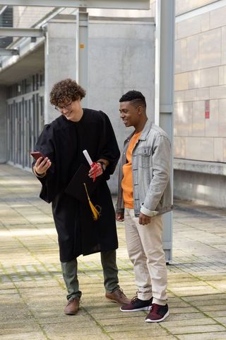 Graduating student showing diploma and phone to friend in university courtyard