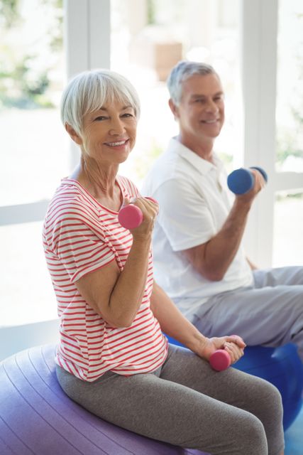 Senior couple sitting on fitness balls, lifting dumbbells, and smiling. Ideal for promoting healthy aging, senior fitness programs, home workout routines, and wellness campaigns targeting older adults.