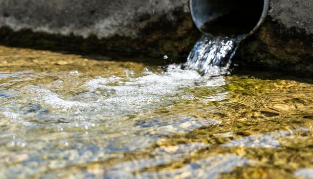 Sunlit water flowing from metal drainage pipe into pebble channel with golden reflections