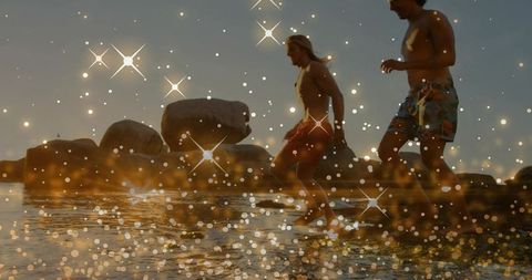 Running and splashing men on rocky shore at sunset with golden bokeh sparkle