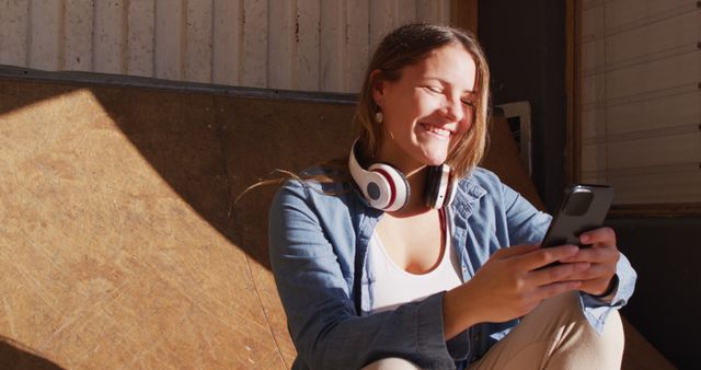 Young woman with headphones around neck enjoying music and texting on smartphone. Sunlight highlights her face as she sits casually, depicting a modern lifestyle that is connected and relaxed. Suitable for promoting technology products, music streaming services, or illustrating leisure and entertainment activities.