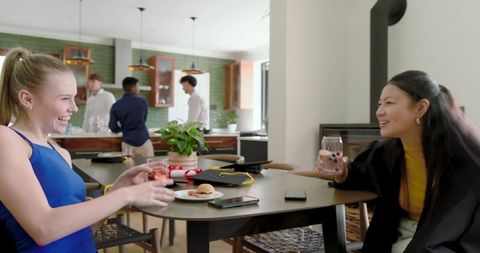 Diverse friends laughing and celebrating graduation at home kitchen table with caps