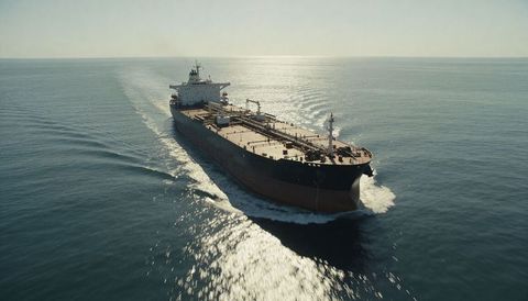 Giant oil tanker cutting bow wave through calm ocean, aerial view of sunlit deck piping