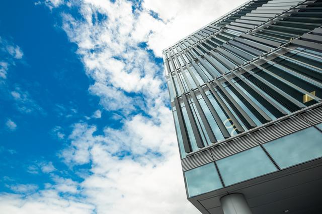 This image showcases a modern office building, focusing on its sleek architectural design and glass facade. The low angle perspective emphasizes the structure against a vibrant blue sky with some cloud formations. Ideal for corporate brochures, real estate advertisements, or articles on urban development and modern construction.