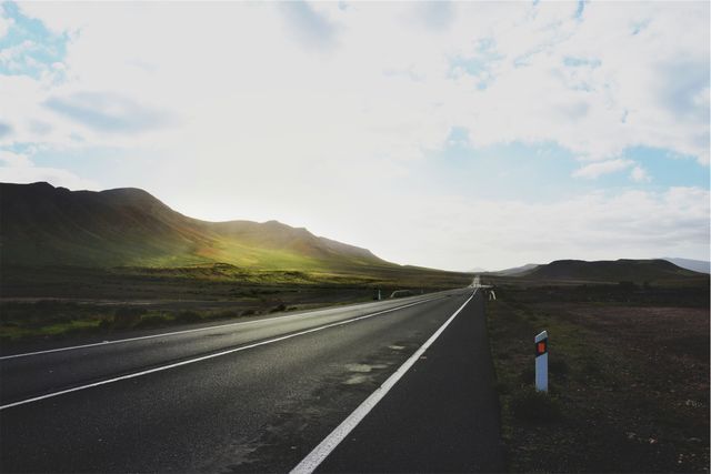 Empty road leading into a tranquil countryside landscape during sunrise. Morning light illuminates the slopes of surrounding hills, creating a serene atmosphere. Ideal for use in travel advertisements, scenic drive promotions, road trip graphics, and nature-themed backgrounds.