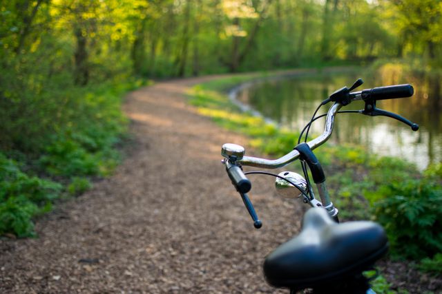 A bicycle is resting on the side of a forest path next to a calm body of water, surrounded by green trees and sunlight filtering through leaves. Perfect for illustrating concepts of cycling, outdoor activities, nature vacations, relaxation, eco-friendly transport, and serene landscapes. Use this for promoting cycling tours, environmental awareness, wellness retreats, or lifestyle blogs emphasizing nature and fitness.