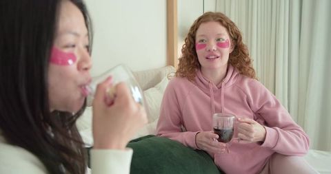 Two friends wearing pink under-eye patches relaxing on bed sipping tea skincare ritual