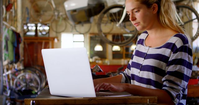 Young woman concentrating on her work while using a laptop in a bicycle workshop. The space is filled with bicycle parts and tools. Suitable for themes of remote work, female entrepreneurship, repair services, and modern technology in traditional workplaces.