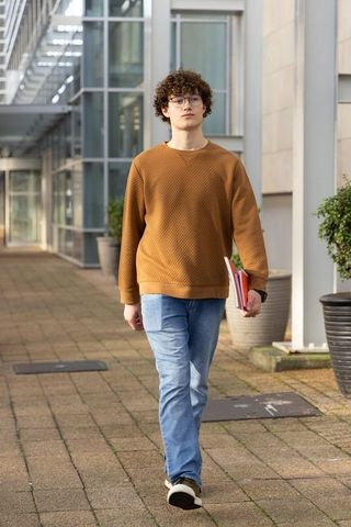 Student walking on modern campus walkway holding notebooks and wearing glasses casual look