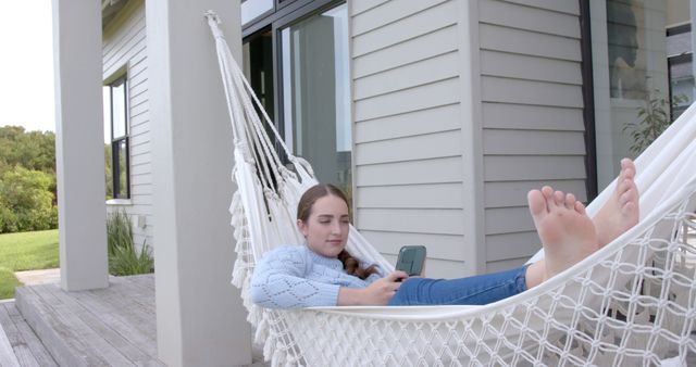 Woman lounging in a hammock on a contemporary patio, looking at her smartphone in leisure wear. Ideal for articles and blog posts about relaxation, modern home living, outdoor spaces, technology, or work-life balance. Also useful for promotional materials for tech gadgets, home and garden products, or lifestyle content.