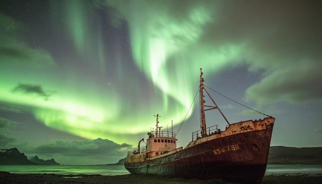 Beached rusted ship under swirling northern lights across remote fjord nightscape with mast