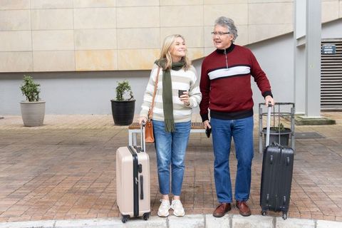 Senior couple traveling with suitcases and coffee outside modern urban building, smiling