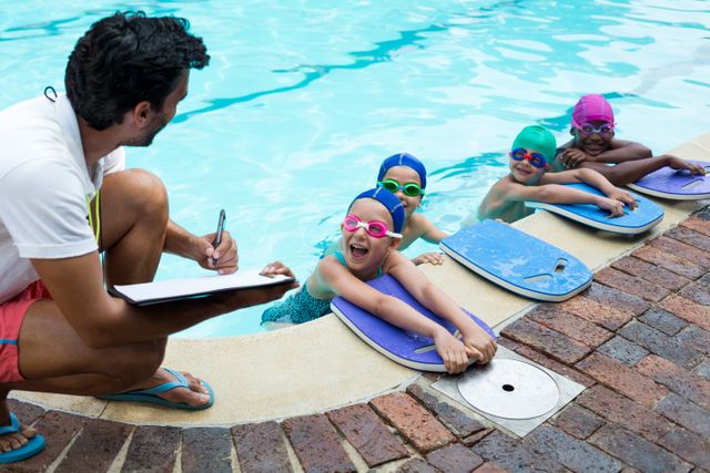 Male instructor coaching young swimmers at poolside. Children wearing swim caps and goggles holding kickboards, smiling and engaging with the coach. Ideal for use in educational materials, swim school promotions, sports training guides, and summer camp advertisements.