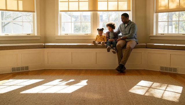 Dad and children reading on sunlit built-in bench with window-pane shadows, cozy home