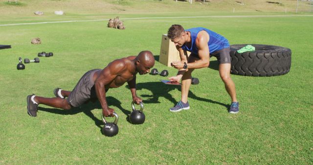 Two diverse fit men exercising outdoors, one doing push ups on kettlebells while the other times him. cross training for fitness at a sports field.