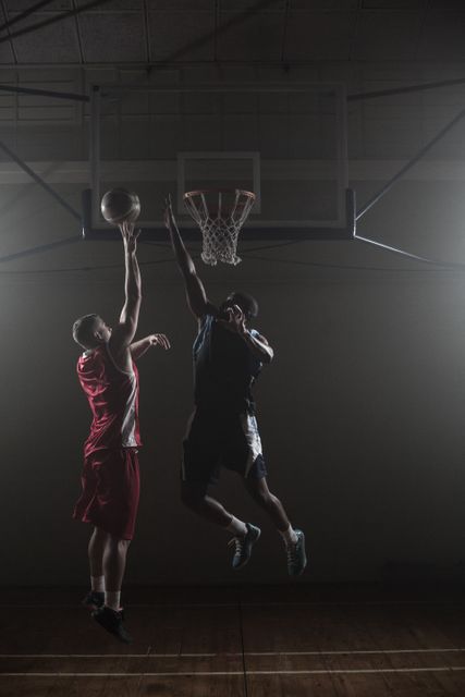 Two basketball players in mid-air, one attempting to score while the other tries to block the shot. Ideal for illustrating sports competition, teamwork, athleticism, and the intensity of basketball games. Suitable for use in sports magazines, promotional materials for basketball events, and fitness-related content.